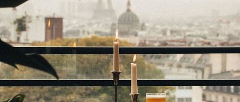 A table with a candle, a glass of amber liquid, and a bowl of food, set against a backdrop of a cityscape with a prominent tower.