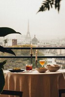 Une table avec une bougie, un verre contenant un liquide ambré et un bol de nourriture, le tout placé devant un paysage urbain où une tour imposante se distingue.
