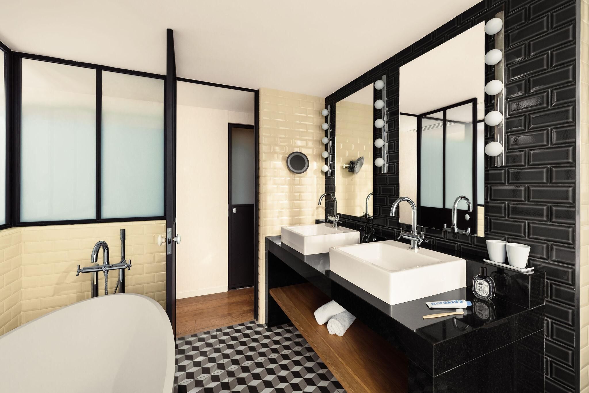 Un baño moderno con piso a cuadros blanco y negro, dos lavamanos blancos, un espejo grande con luces y una bañera.