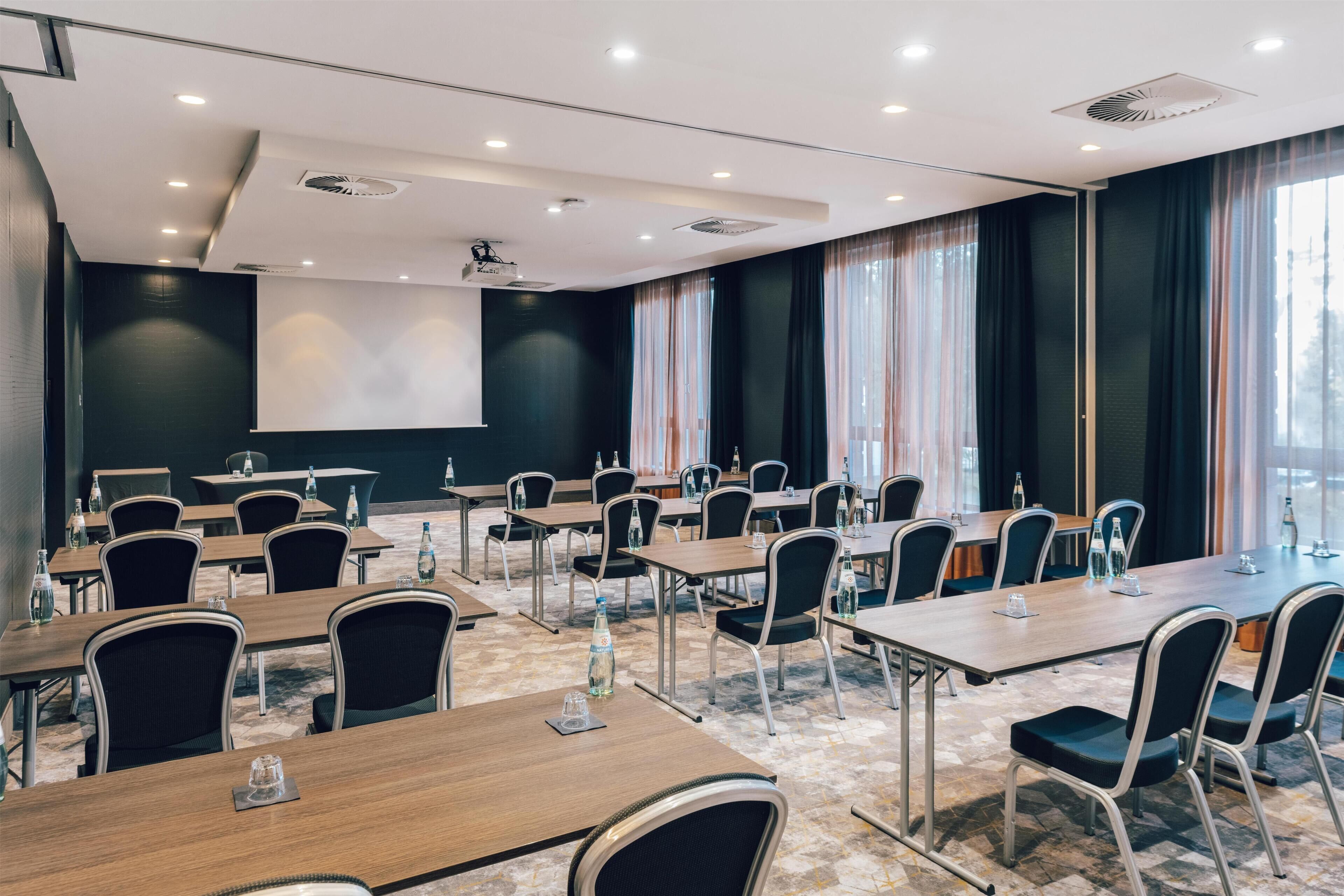 A conference room with multiple tables and chairs, a projector screen, and large windows with curtains.