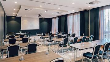 Ein Konferenzraum mit mehreren Tischen und Stühlen, einer Projektionsleinwand und großen Fenstern mit Vorhängen.