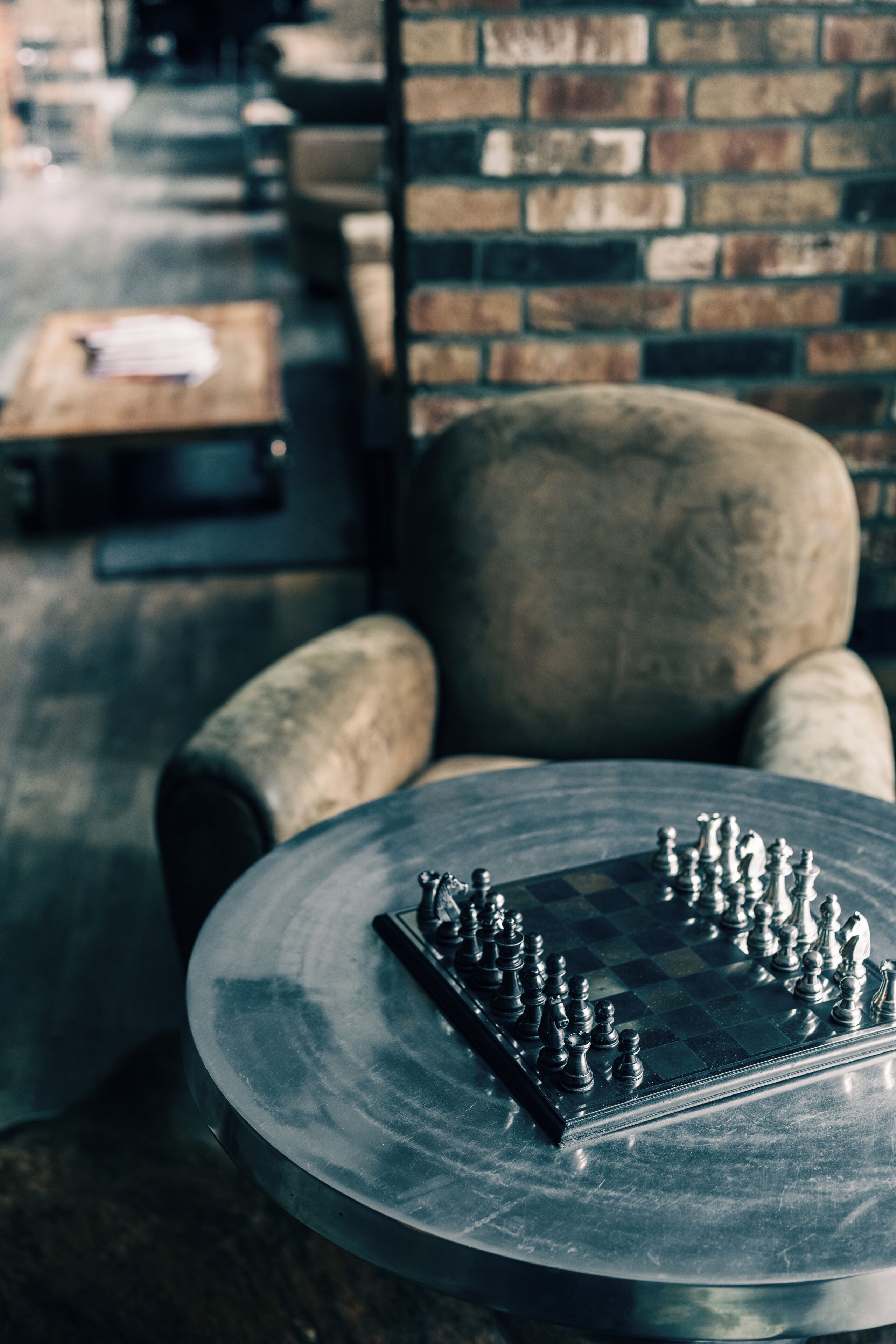 Een ronde metalen tafel met een schaakbord in een kamer met bakstenen muren en een leren fauteuil.
