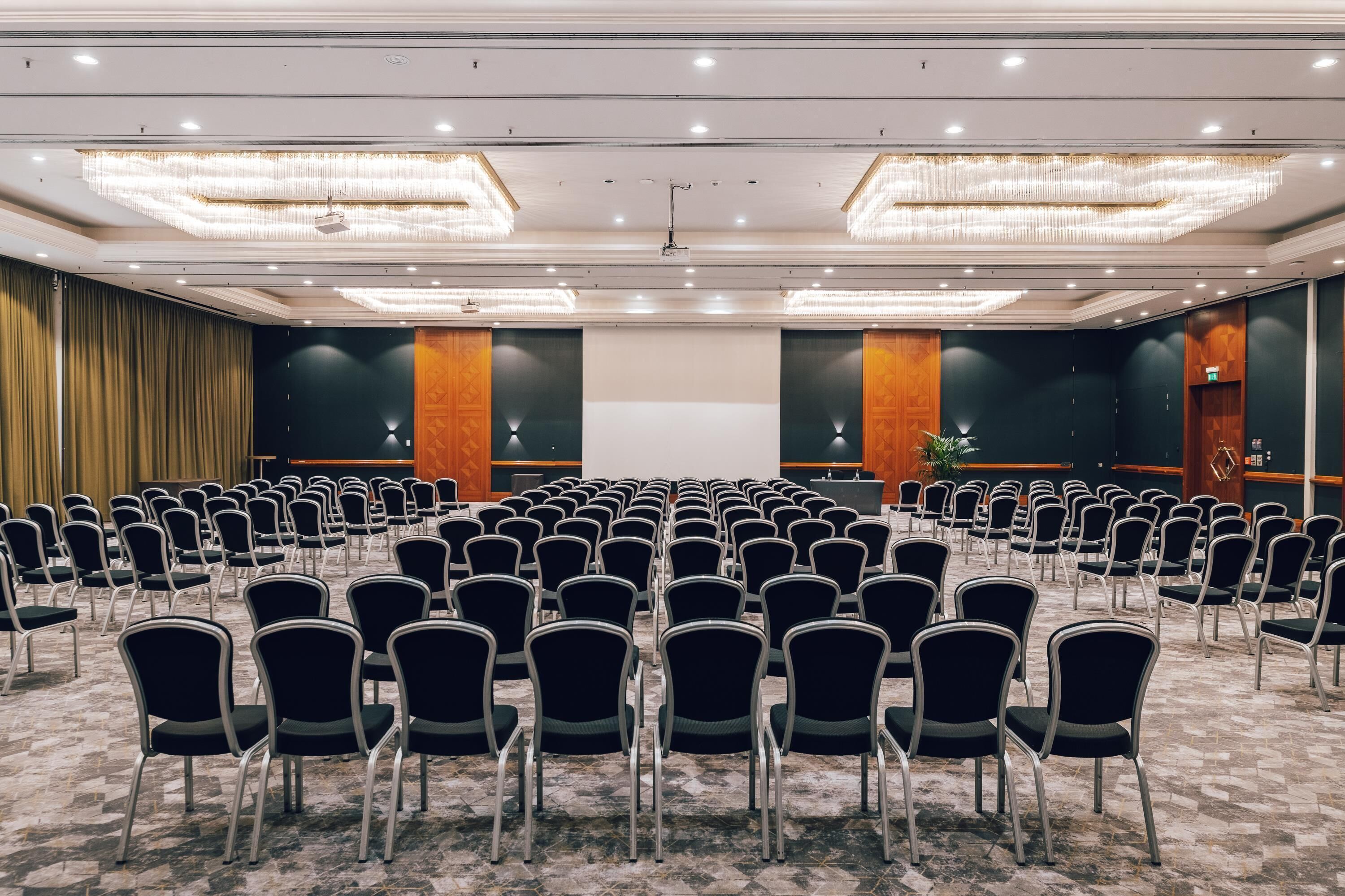 Un salón de conferencias con filas de sillas, una pantalla de proyector y un escenario.