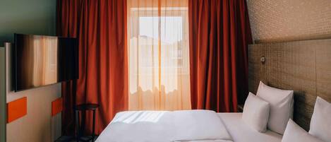 Hotellihuone, jossa on sänky, televisio ja ikkuna, jossa on verhot.