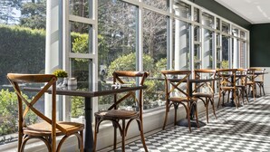 Une pièce lumineuse, baignée de soleil, avec de grandes fenêtres, un sol à carreaux et des chaises en bois dotées d’assises en osier tressé.