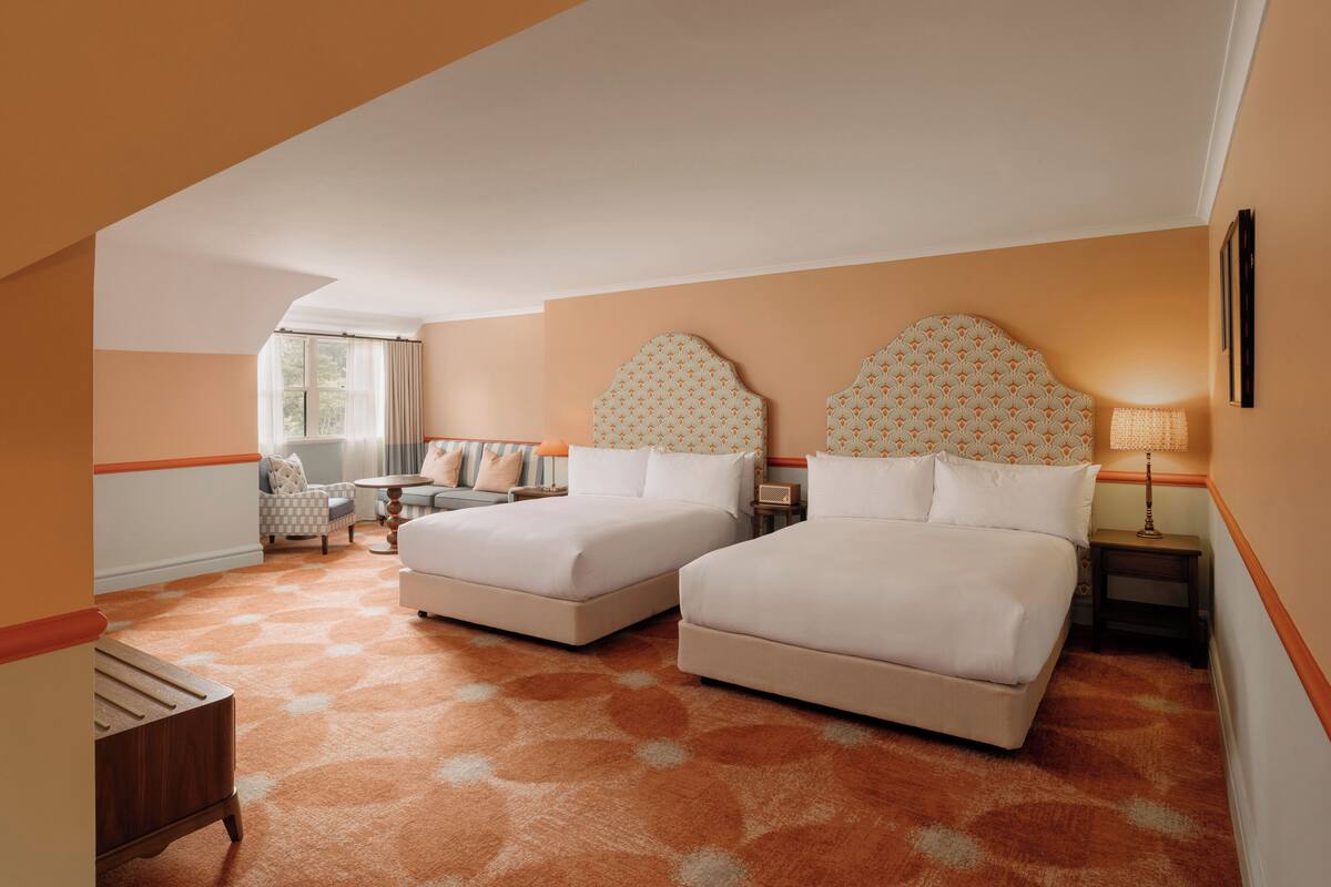 A hotel room with two beds, a small table, a chair, and a window with curtains.
