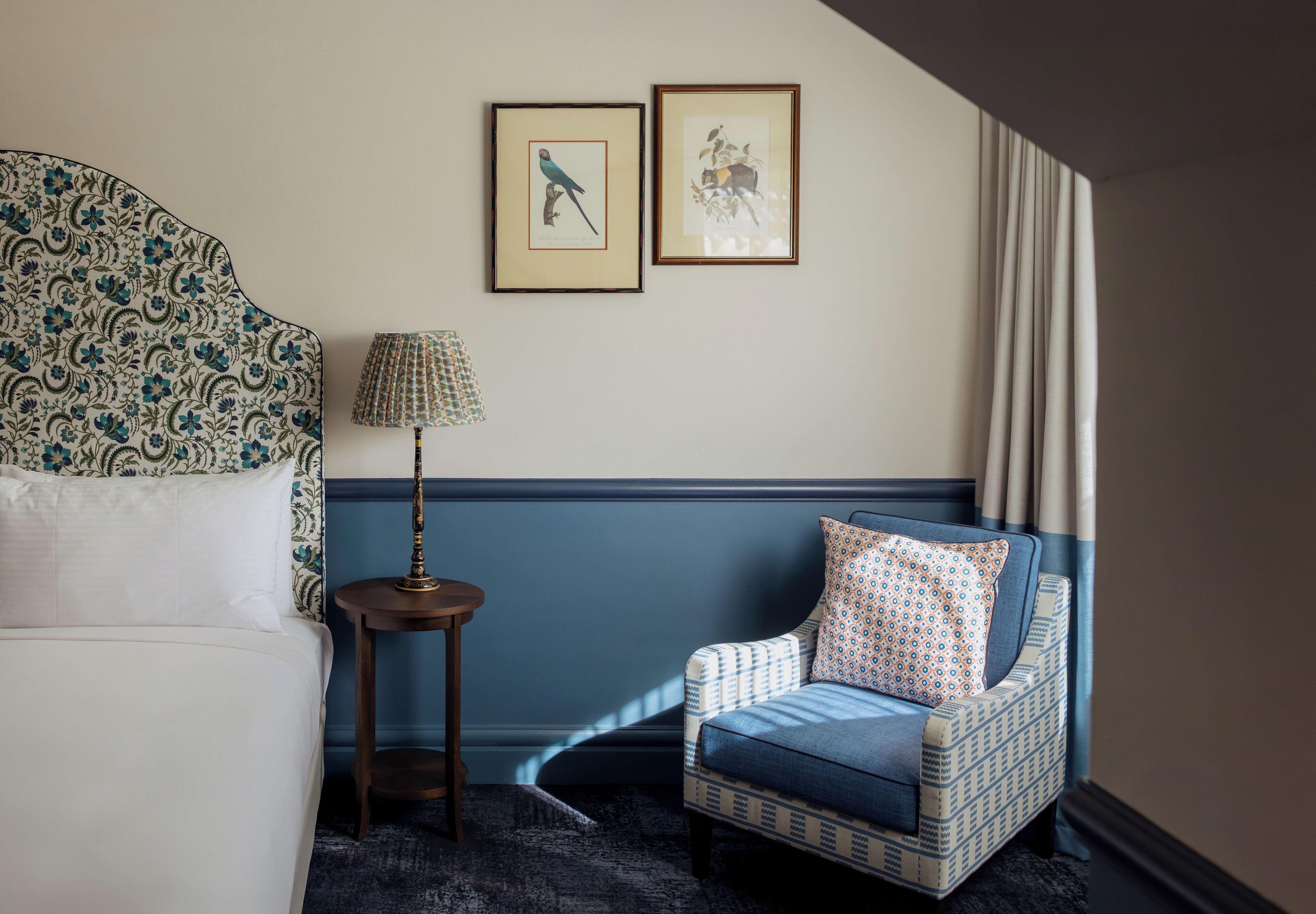 Ein Zimmer mit einem Bett, einem verzierten Kopfteil, einem blauen Sessel mit einem gemusterten Kissen, einem kleinen Tisch mit einer Lampe sowie zwei gerahmten Bildern an der Wand.