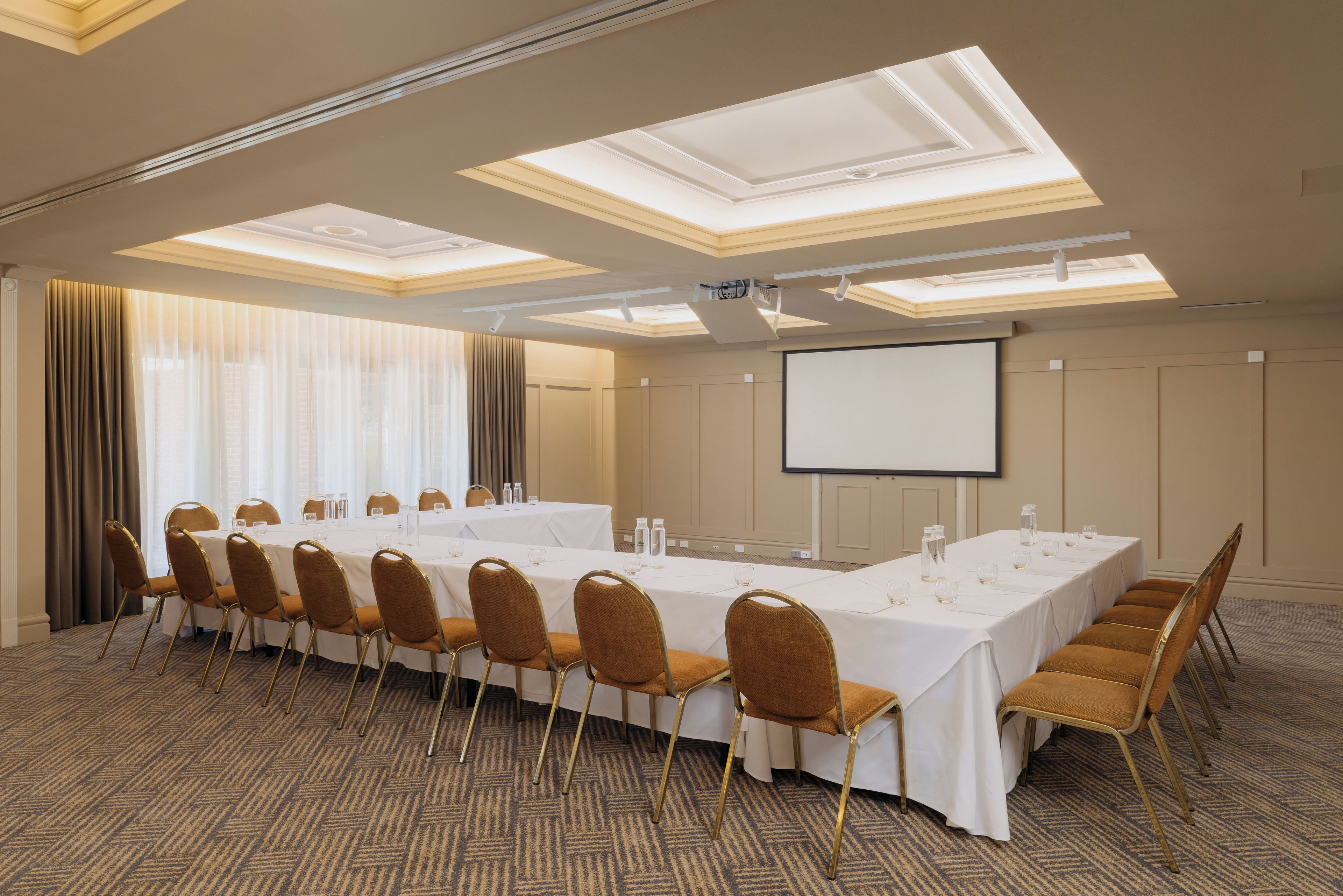 Une salle de conférence avec une longue table, des chaises, un écran de projection et une grande fenêtre avec des rideaux.