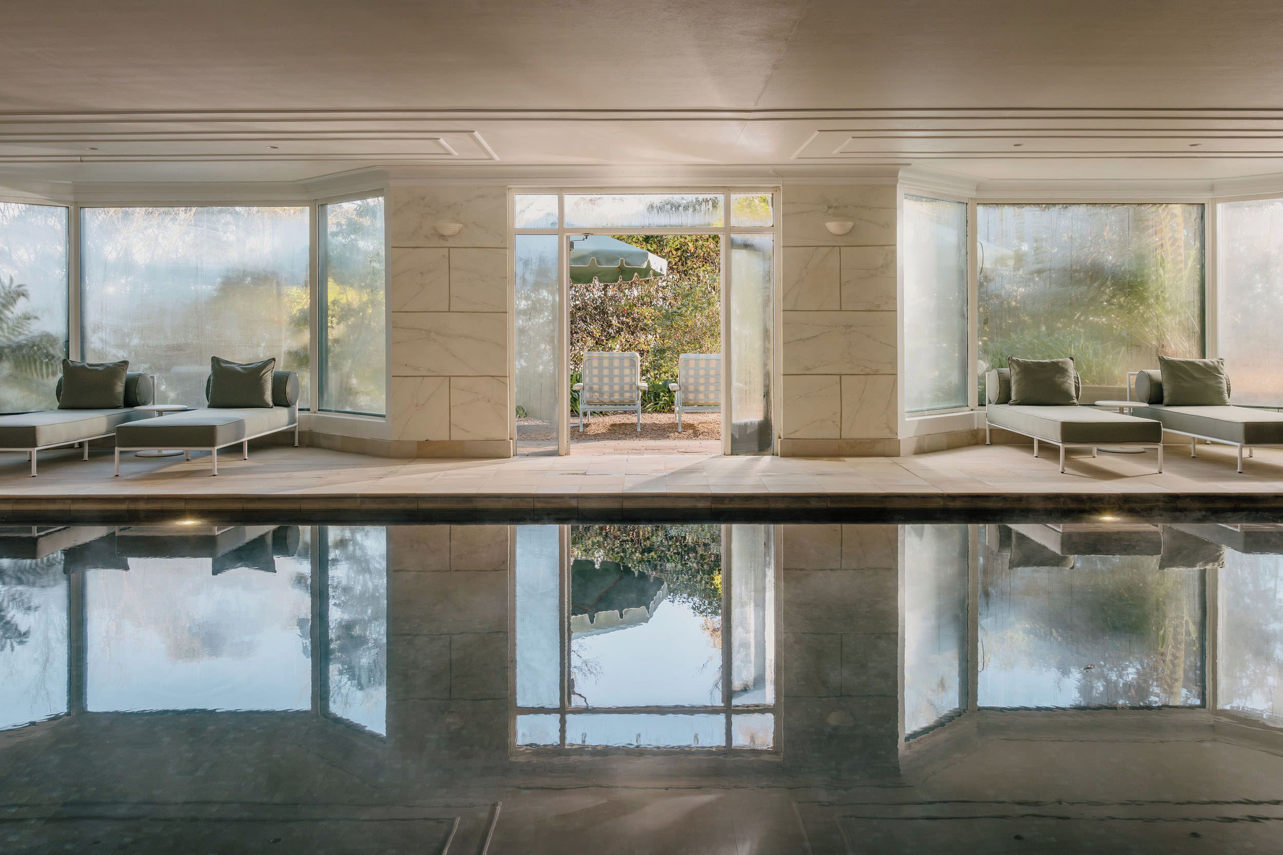 A modern indoor pool area with clear glass walls, seating on both sides, and a view of a garden.