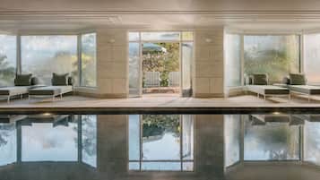 A modern indoor pool area with clear glass walls, seating on both sides, and a view of a garden.
