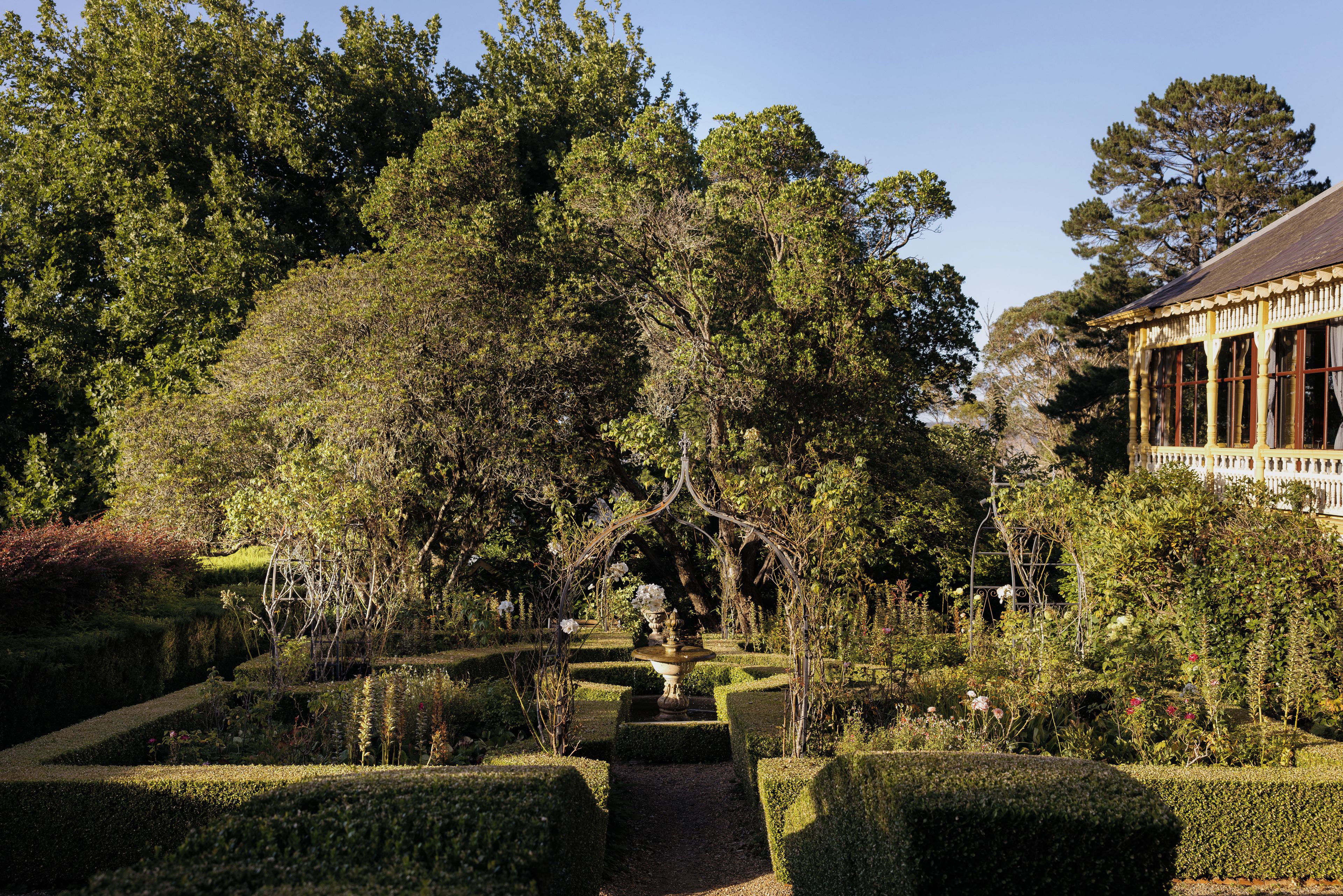 Un jardín formal con setos recortados, una fuente central y un edificio clásico con ventanas arqueadas.