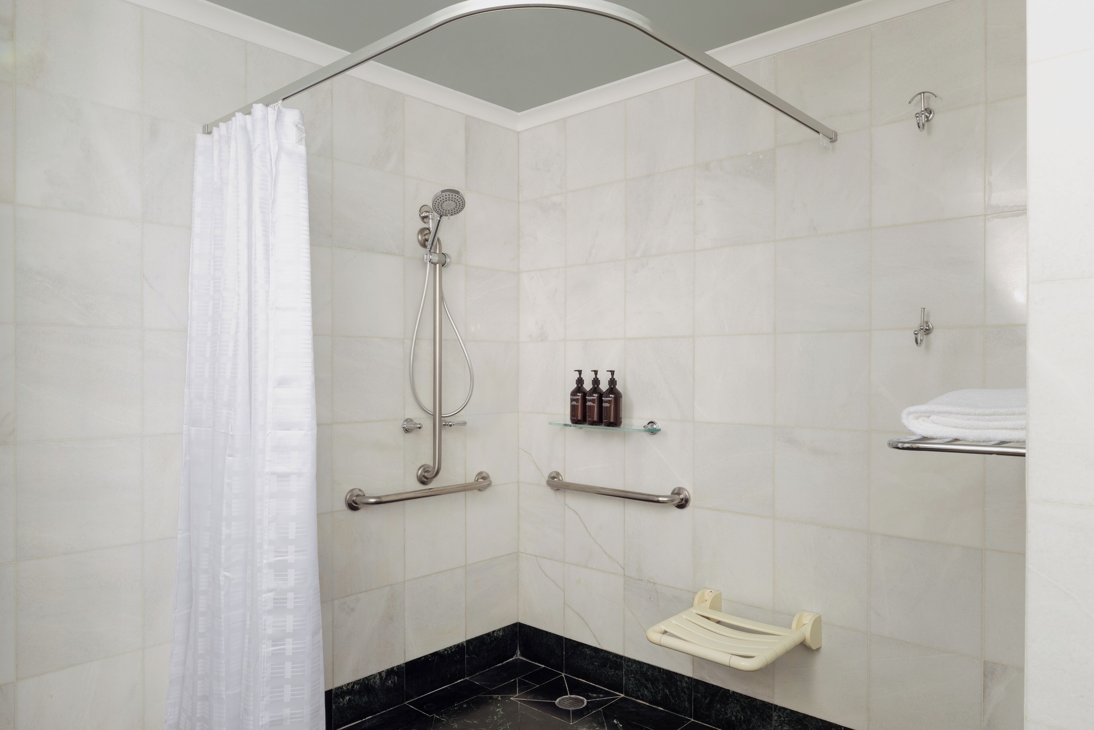 A bathroom with a shower, grab bars, a towel rack, and a soap dispenser.