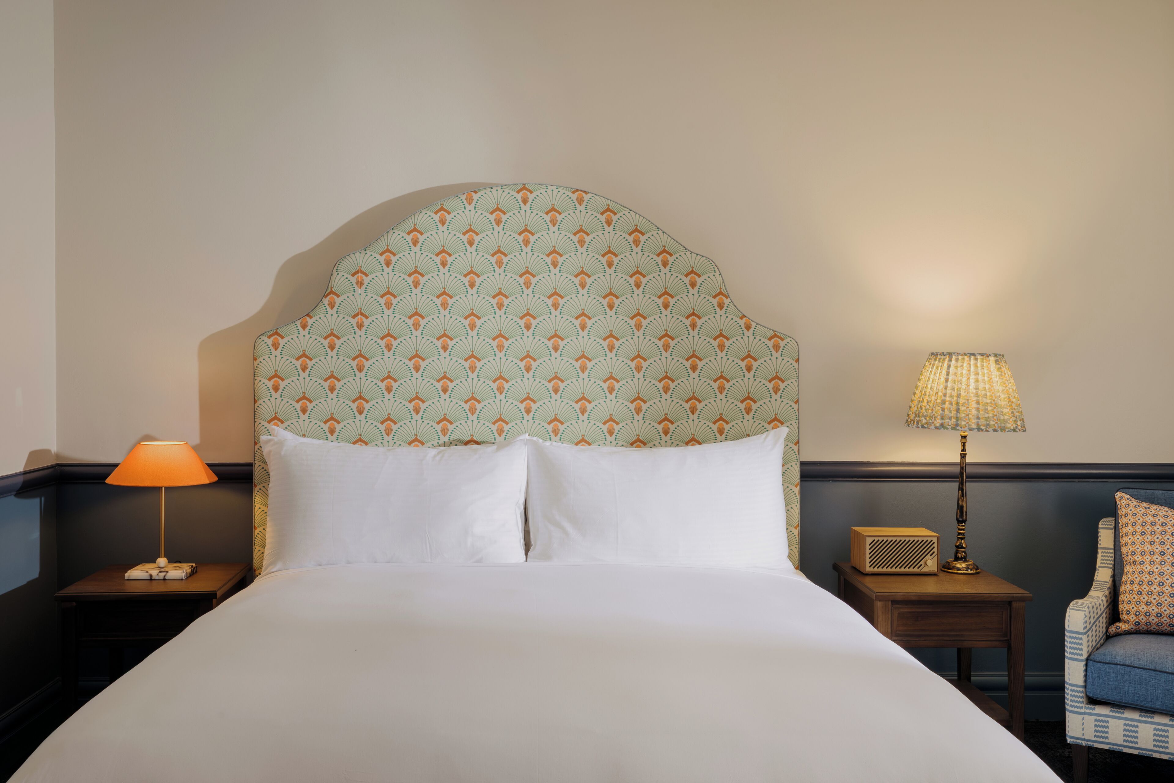 A neatly made bed with a patterned headboard, a bedside lamp, a radio, and a chair with a cushion.