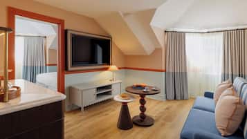 A modern hotel room with a TV mounted on the wall, a sofa, a small table with a lamp, and a dining area with a tray and a plate.