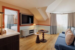 A modern hotel room with a TV mounted on the wall, a sofa, a small table with a lamp, and a dining area with a tray and a plate.