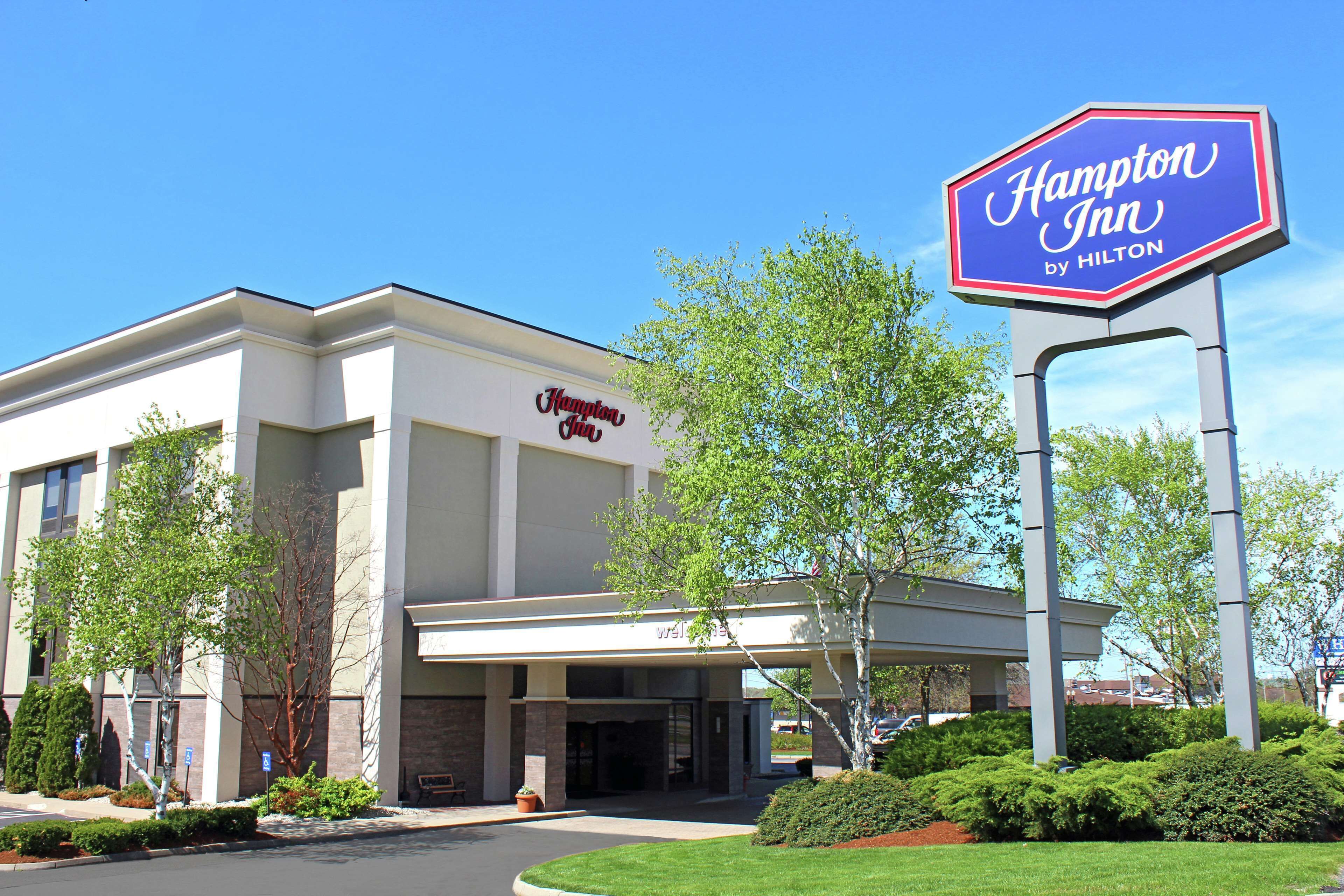 A Hampton Inn by Hilton hotel with a clear entrance, a signboard, and a well-maintained lawn.