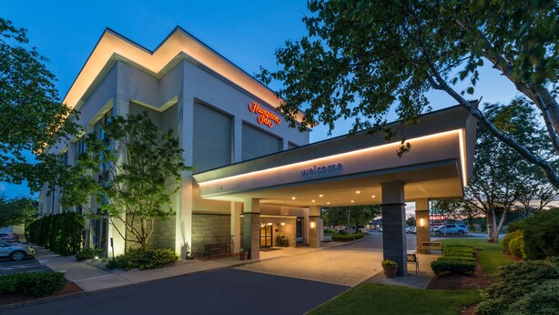 A modern hotel entrance with a 'Welcome' sign, a covered driveway, and a parking area.