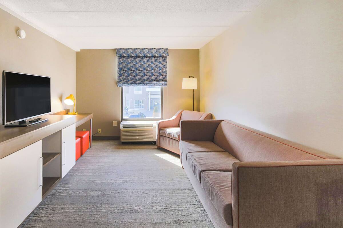 Hotellihuone, jossa on taulutelevisio, sohva, pieni pöytä lampun kanssa ja ikkuna, josta on näkymä rakennuksiin.