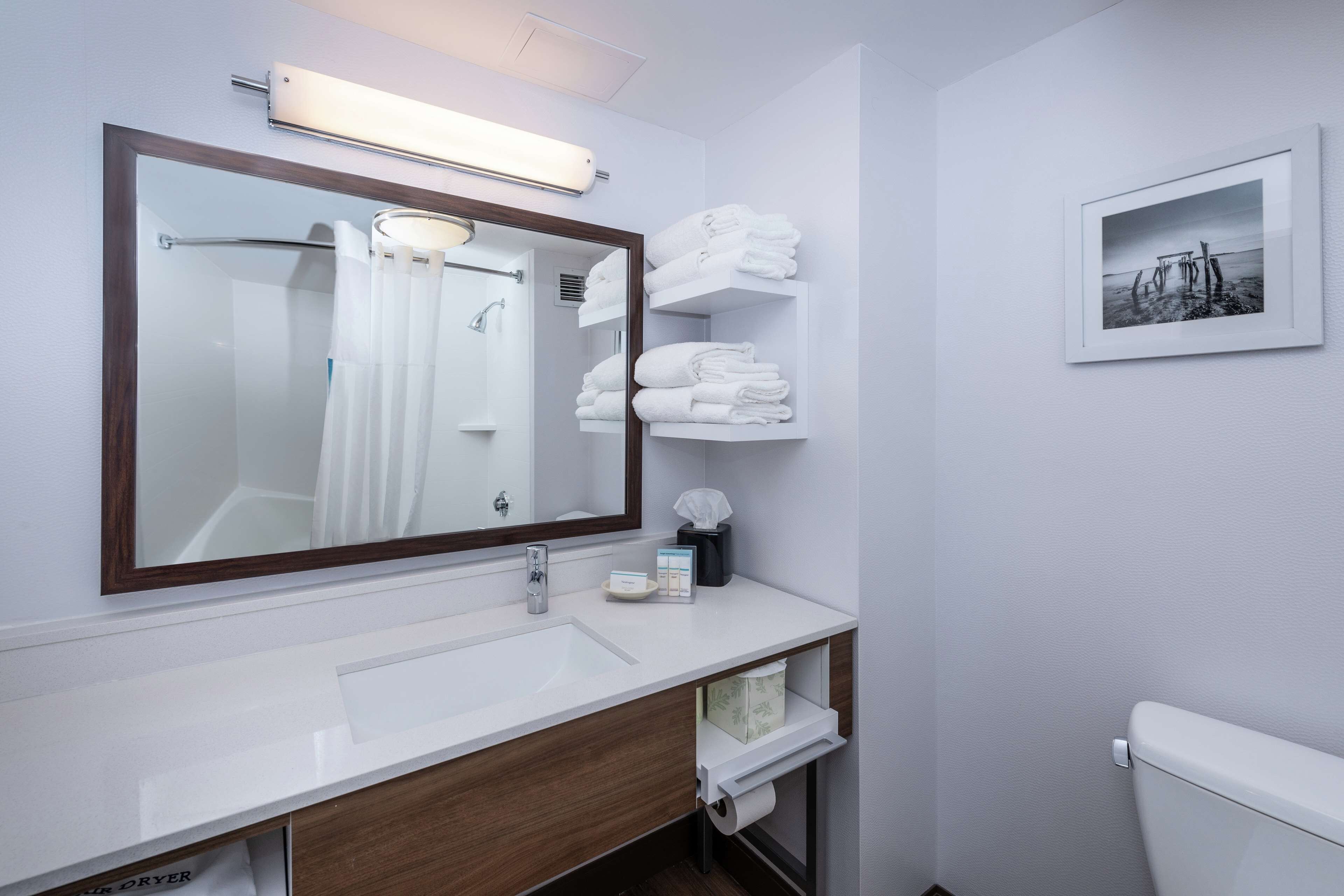 A modern bathroom with a large mirror, white sink, and a toilet. There are towels stacked on a shelf and a framed picture on the wall.