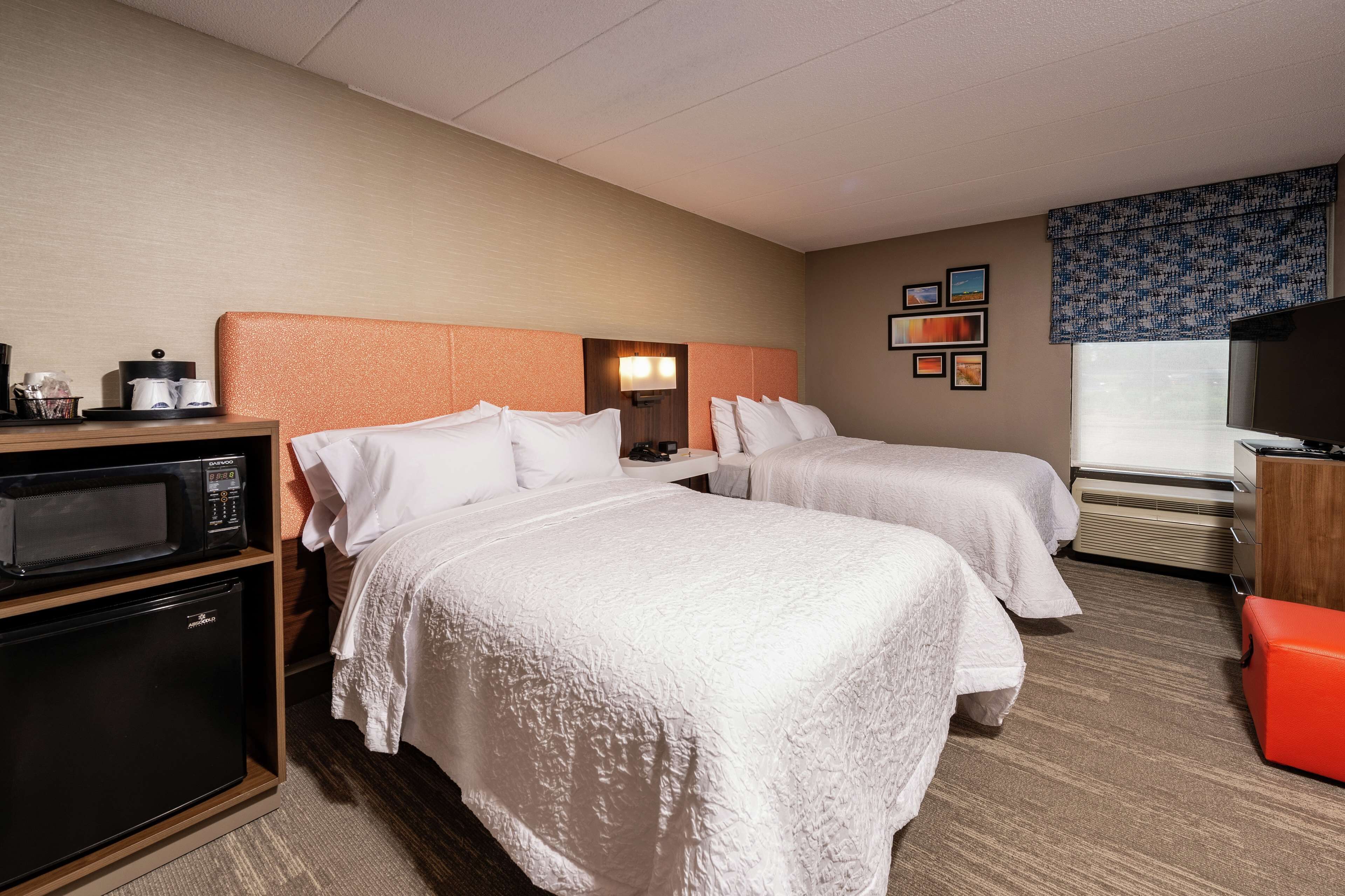 A hotel room with two beds, a microwave, a mini fridge, a TV, and a red ottoman.