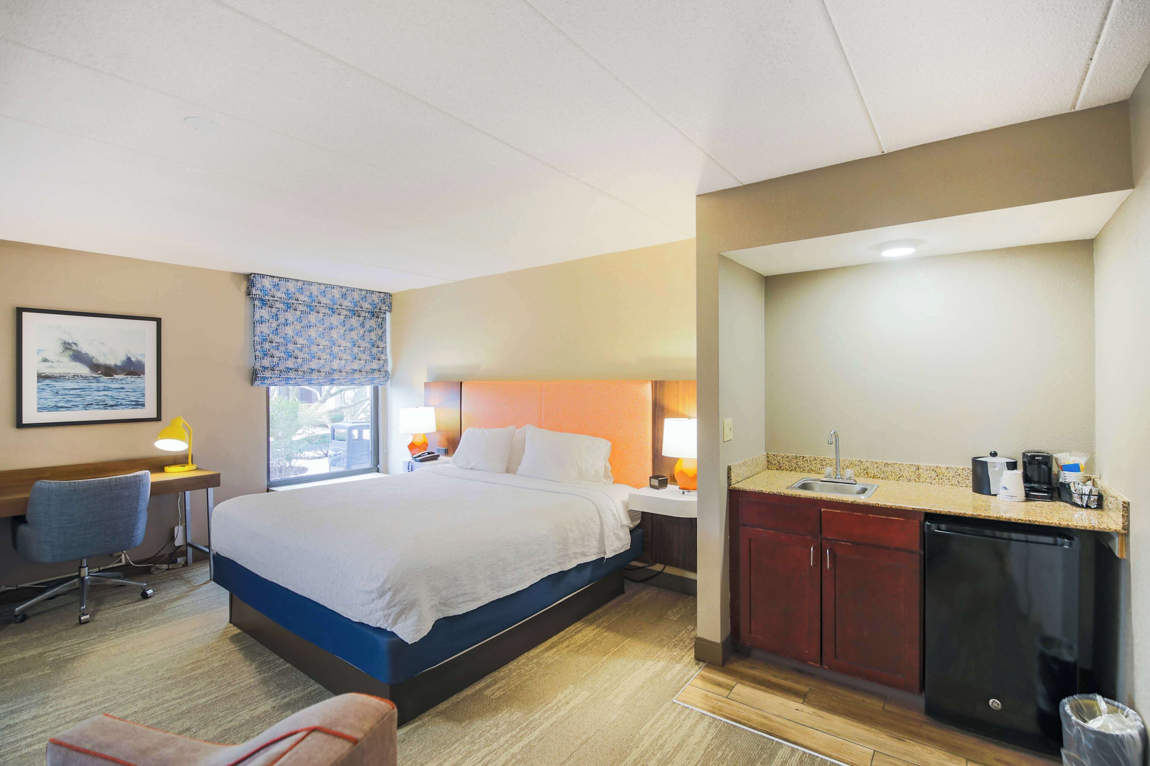 A hotel room with a large bed, a desk with a chair, a small kitchenette, and a window with a view.