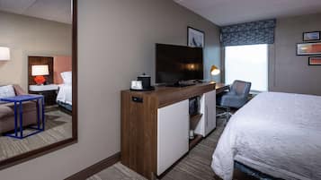 A hotel room with a bed, a desk with a television, a chair, a mirror, and a window with a view.