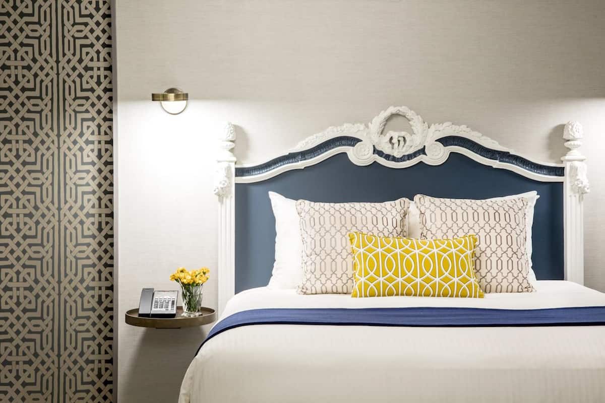 A neatly made bed with a white and blue headboard, two patterned pillows, and a bedside table with a phone and flowers.