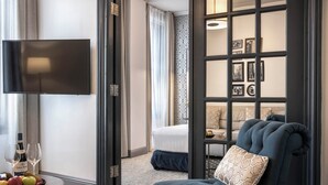 A modern hotel room with a flat-screen TV, a dining table with fruit and wine, a comfortable sofa, and a bed with a view.