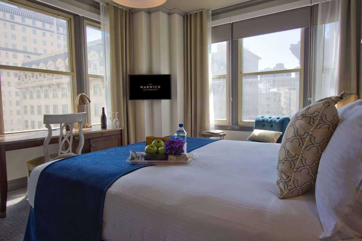 A hotel room with a large bed, a flat-screen TV on the wall, a desk with a chair, and a window with a city view.