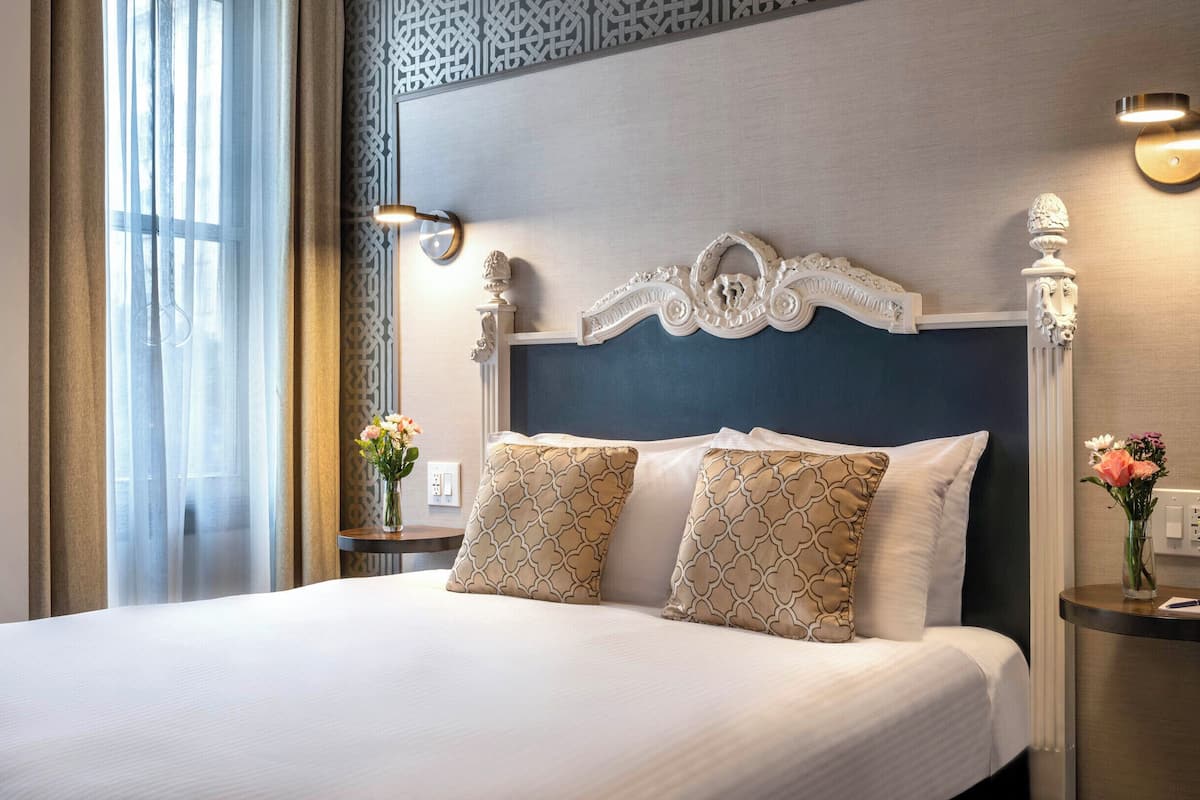 A hotel room with a large bed, decorative headboard, bedside tables with lamps, and a view of the outdoors.