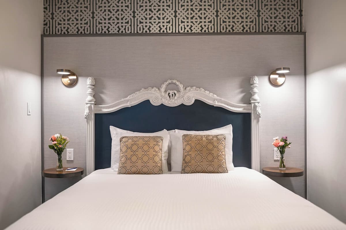 A hotel room with a large bed, two bedside tables with vases, a decorative headboard, and a patterned wall panel.
