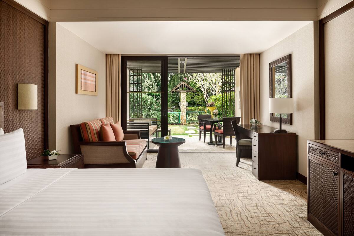 A hotel room with a large bed, a sofa, a desk, and a dining area with a view of the garden.