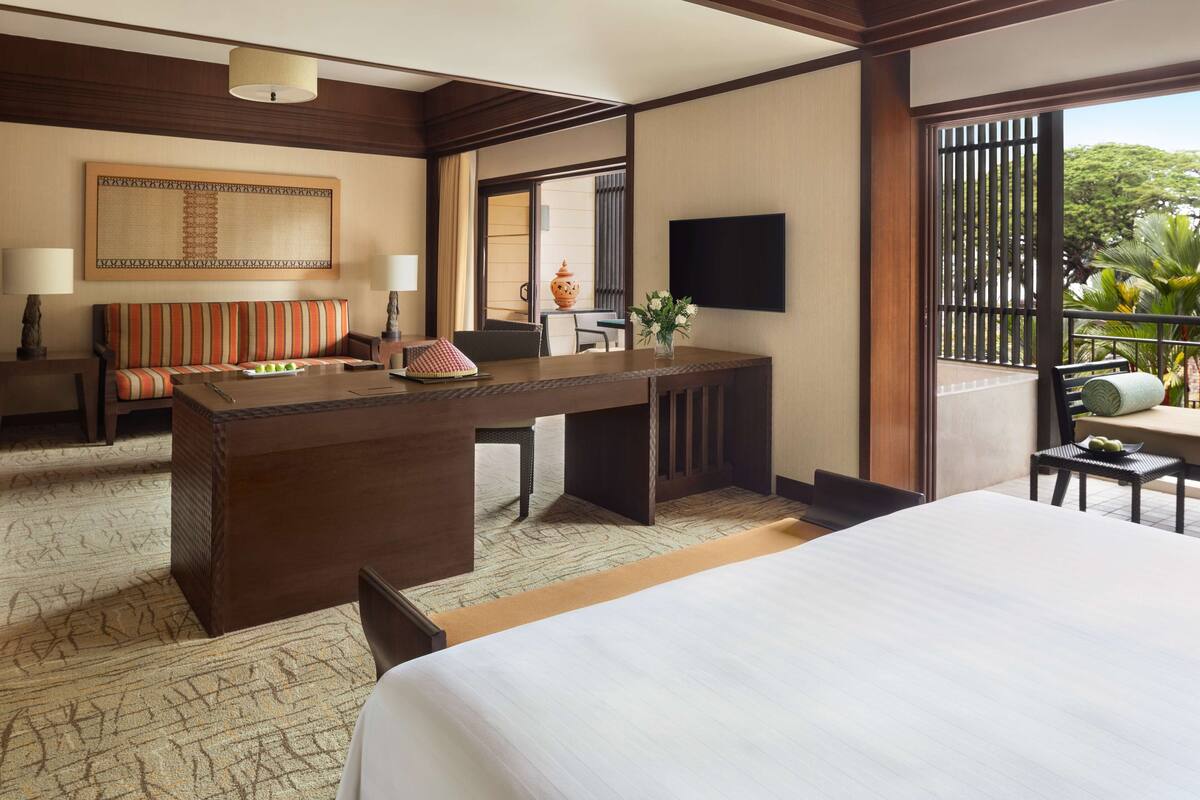 A hotel room with a bed, a desk with a chair, a sofa, a TV, and a balcony with plants.