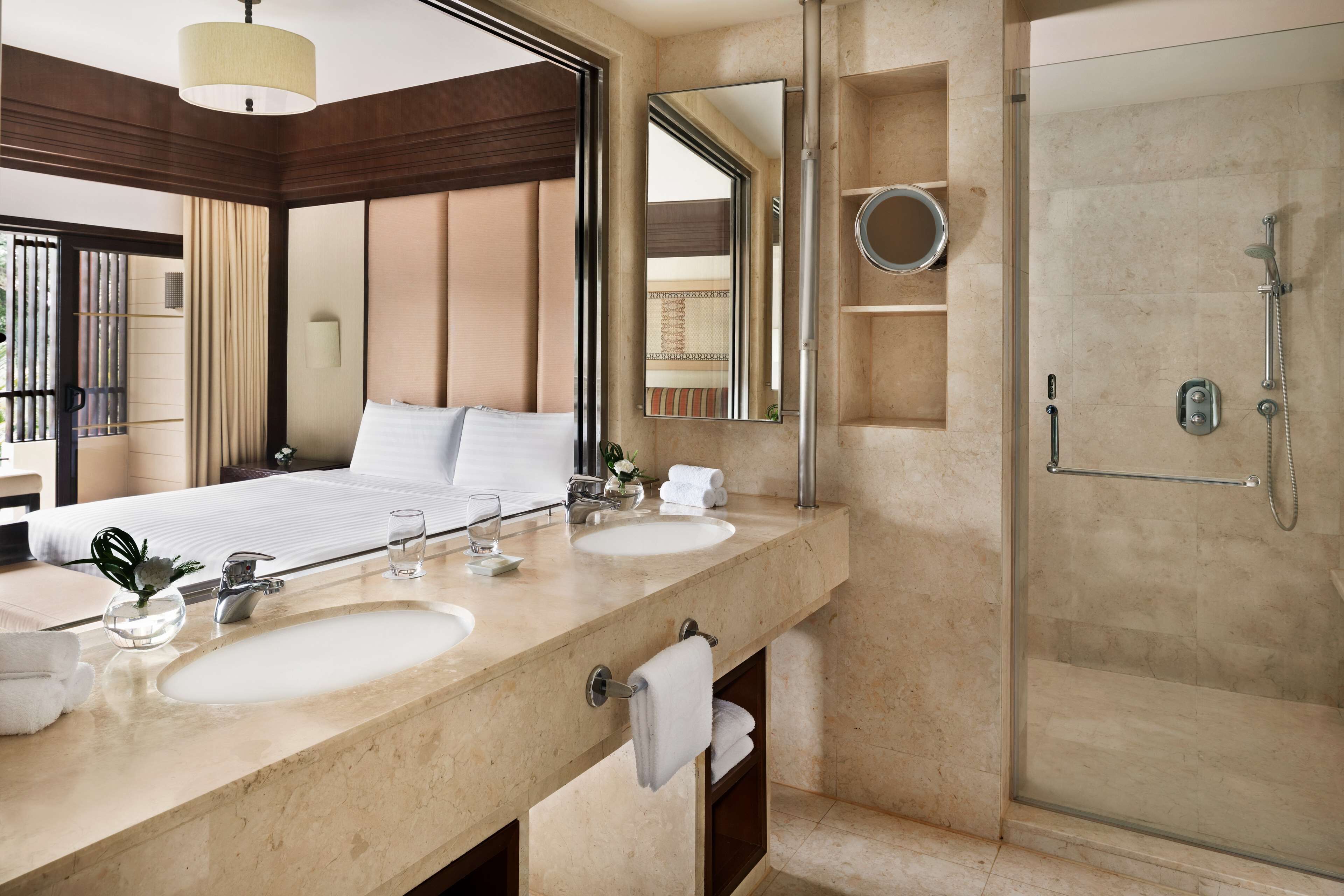 A hotel bathroom with a large mirror, two sinks, a shower, and a bed with a view of the outdoors.