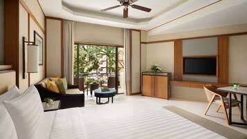 A hotel room with a large bed, a ceiling fan, a TV, a dining table, and a view of greenery outside.