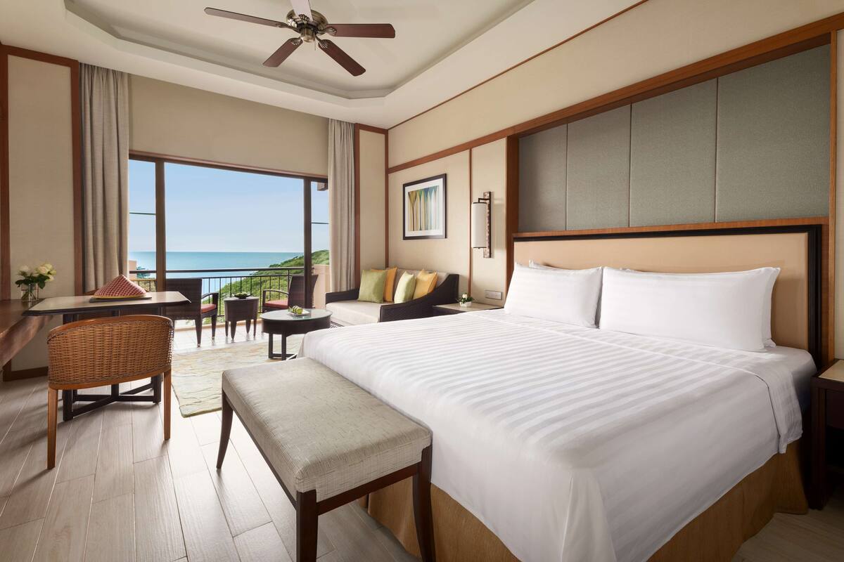 A hotel room with a large bed, a desk, a chair, a small table, and a balcony with a view of the sea.