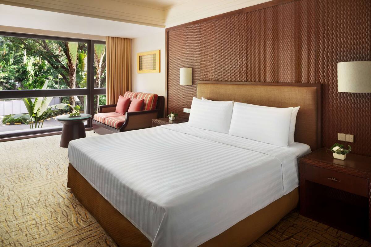 A large bed with white linens, a wooden headboard, and a nightstand with a lamp. There is a window with a view of greenery outside.