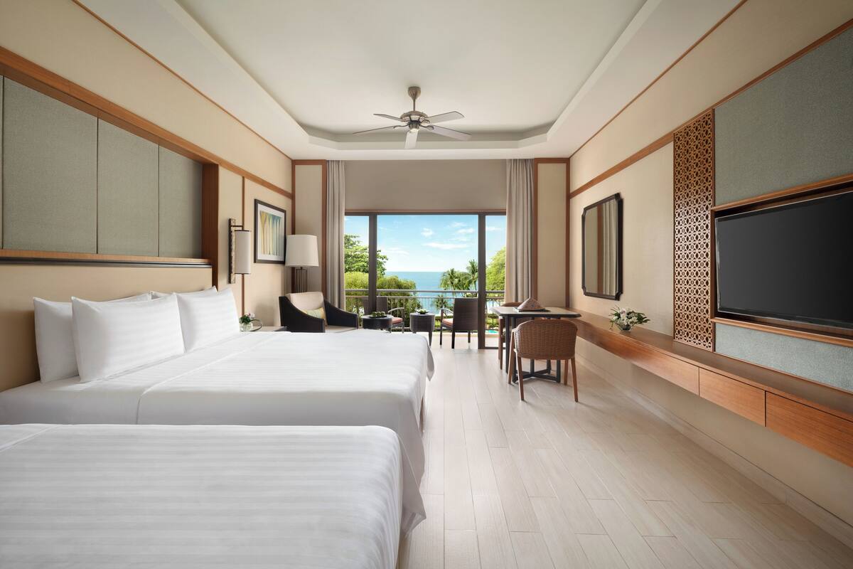 A hotel room with two large beds, a ceiling fan, a TV, a dining area with a table and chairs, and a view of the outdoors.