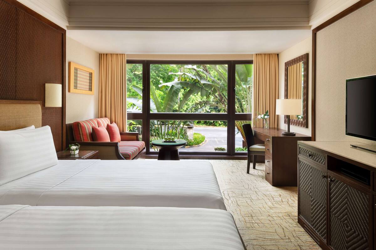 A hotel room with a large bed, a desk, a chair, a television, and a view of greenery outside.