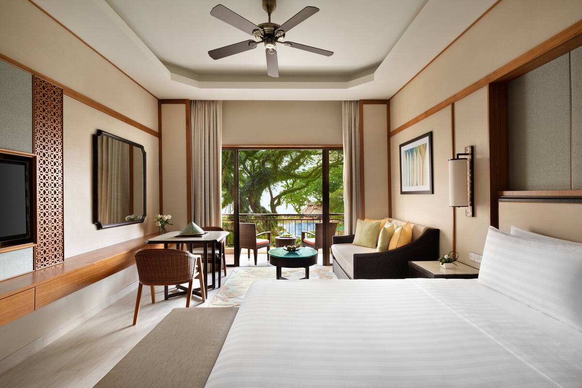 A hotel room with a bed, a desk, a chair, a sofa, a ceiling fan, and a window with a view of trees.