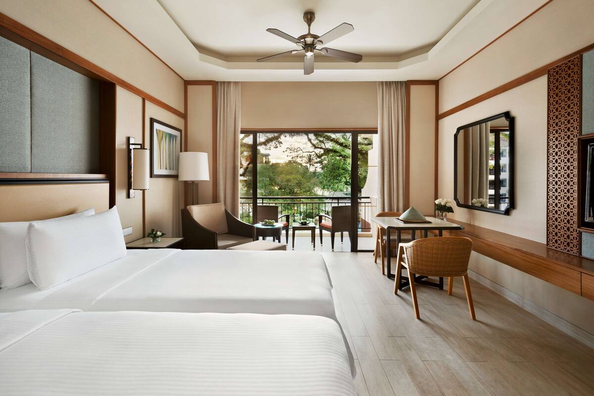 A hotel room with two beds, a desk, a chair, a balcony with a view of trees, and a ceiling fan.