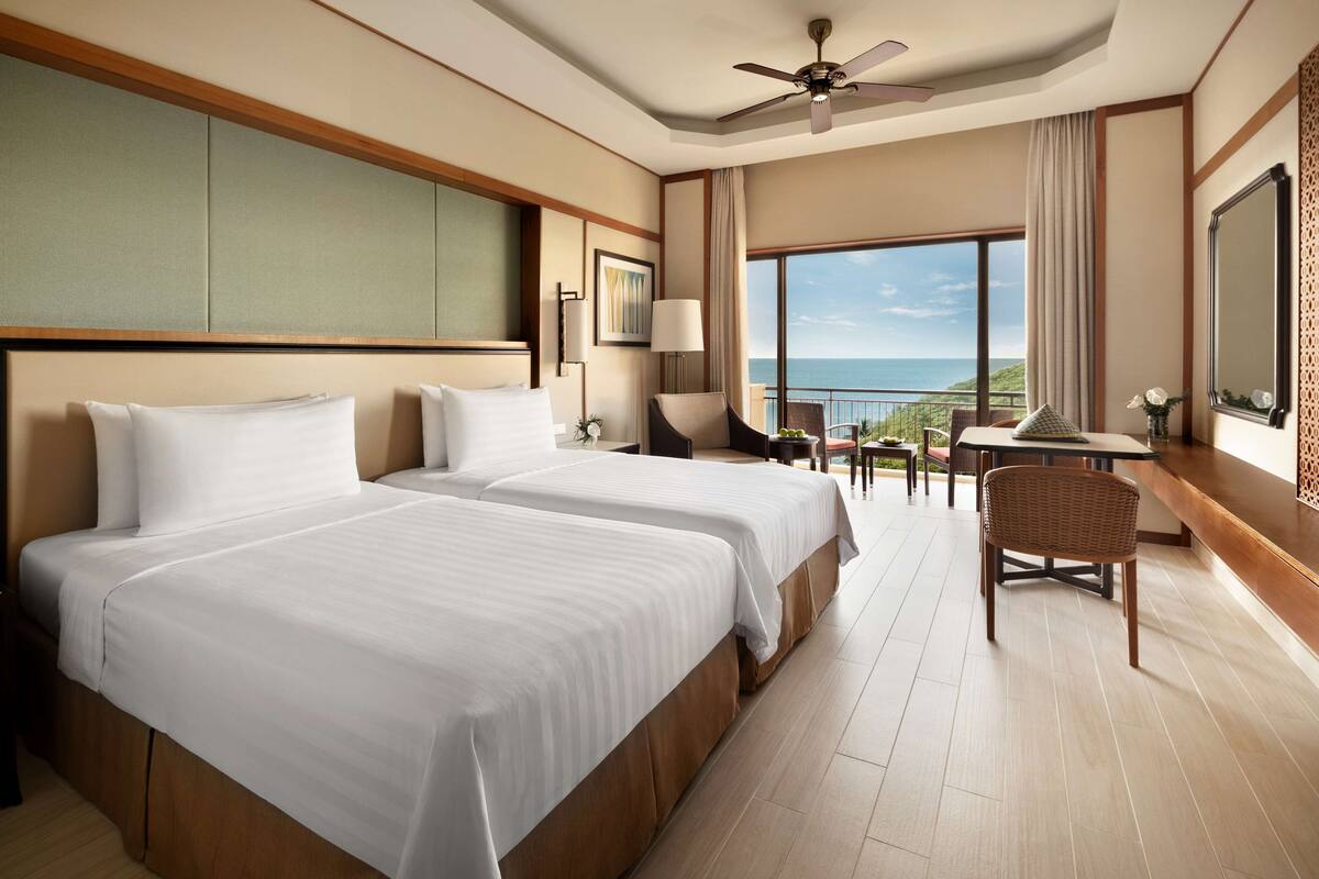 A hotel room with two beds, a balcony with a view of the sea, a ceiling fan, and a dining area.