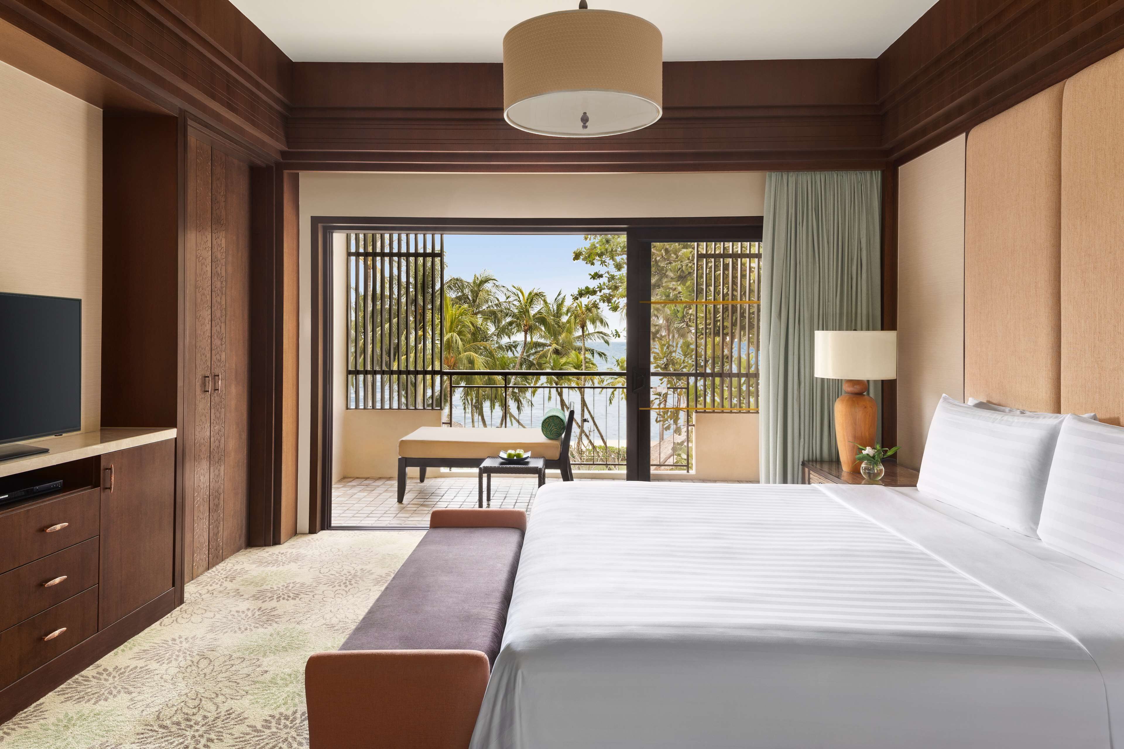 A hotel room with a large bed, a TV, a desk, and a balcony with a view of palm trees and the sea.