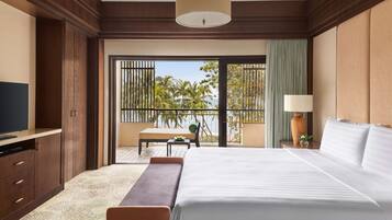 A hotel room with a large bed, a TV, a desk, and a balcony with a view of palm trees and the sea.