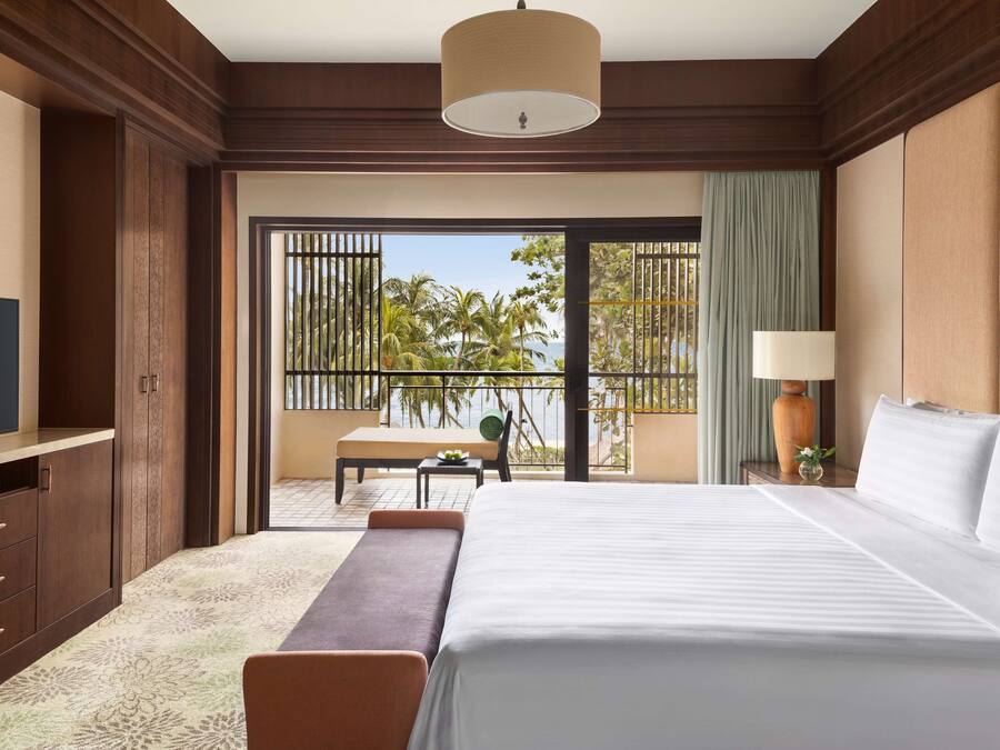 A hotel room with a large bed, a TV, a desk, and a balcony with a view of palm trees and the sea.