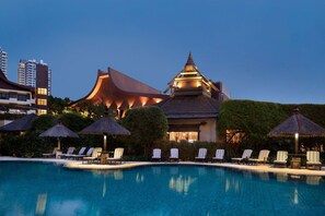 A swimming pool with lounge chairs and umbrellas, surrounded by well-maintained greenery and a modern building with traditional architectural elements.