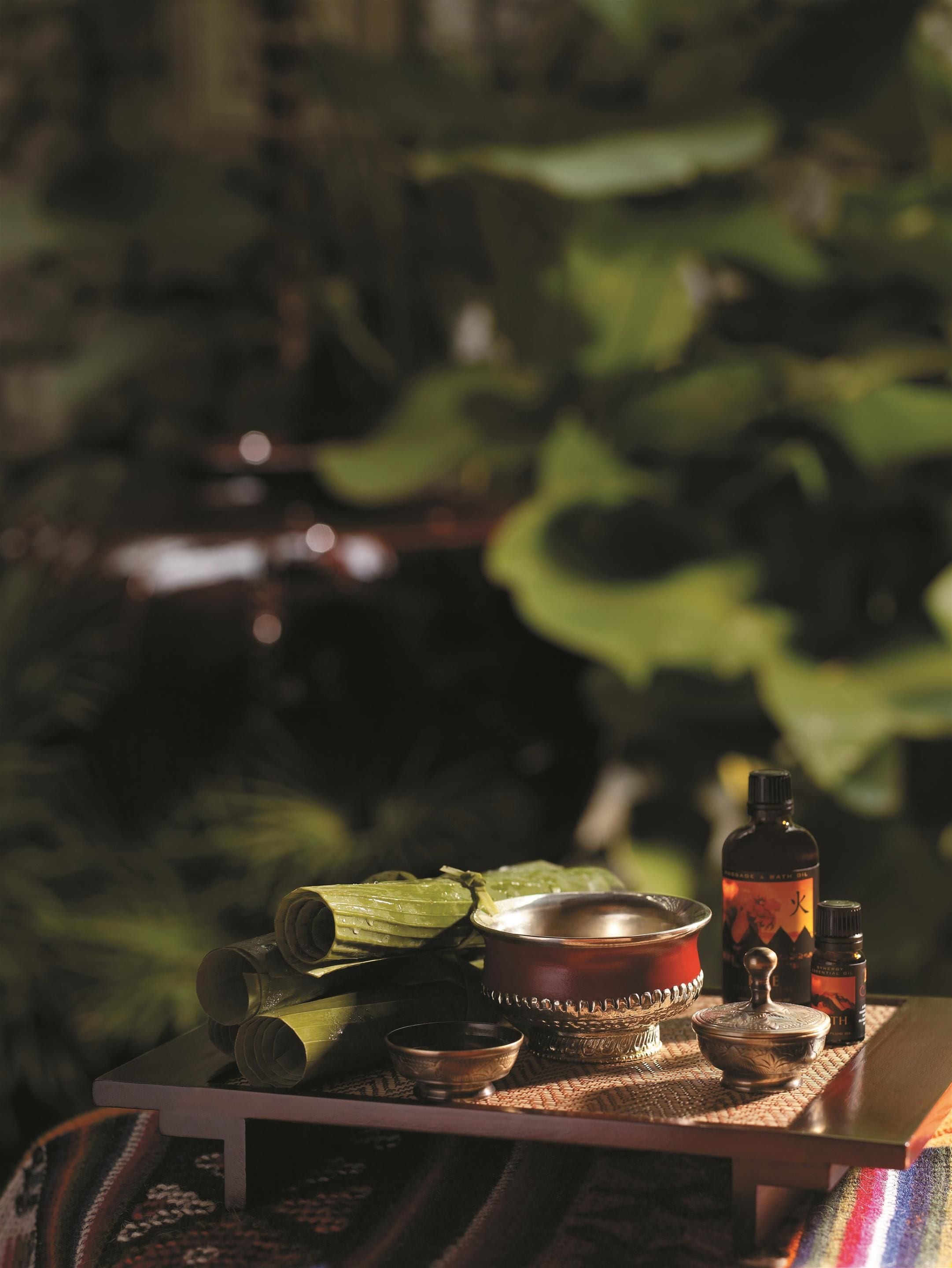 A table with traditional items including a metal bowl, a bottle, and rolled-up mats, set against a backdrop of greenery.