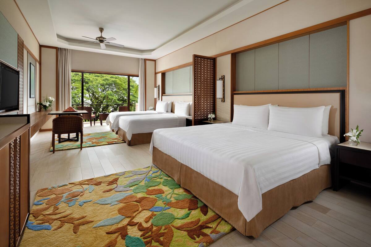 A hotel room with a large bed, a desk, a chair, a TV, and a balcony with a view of trees.