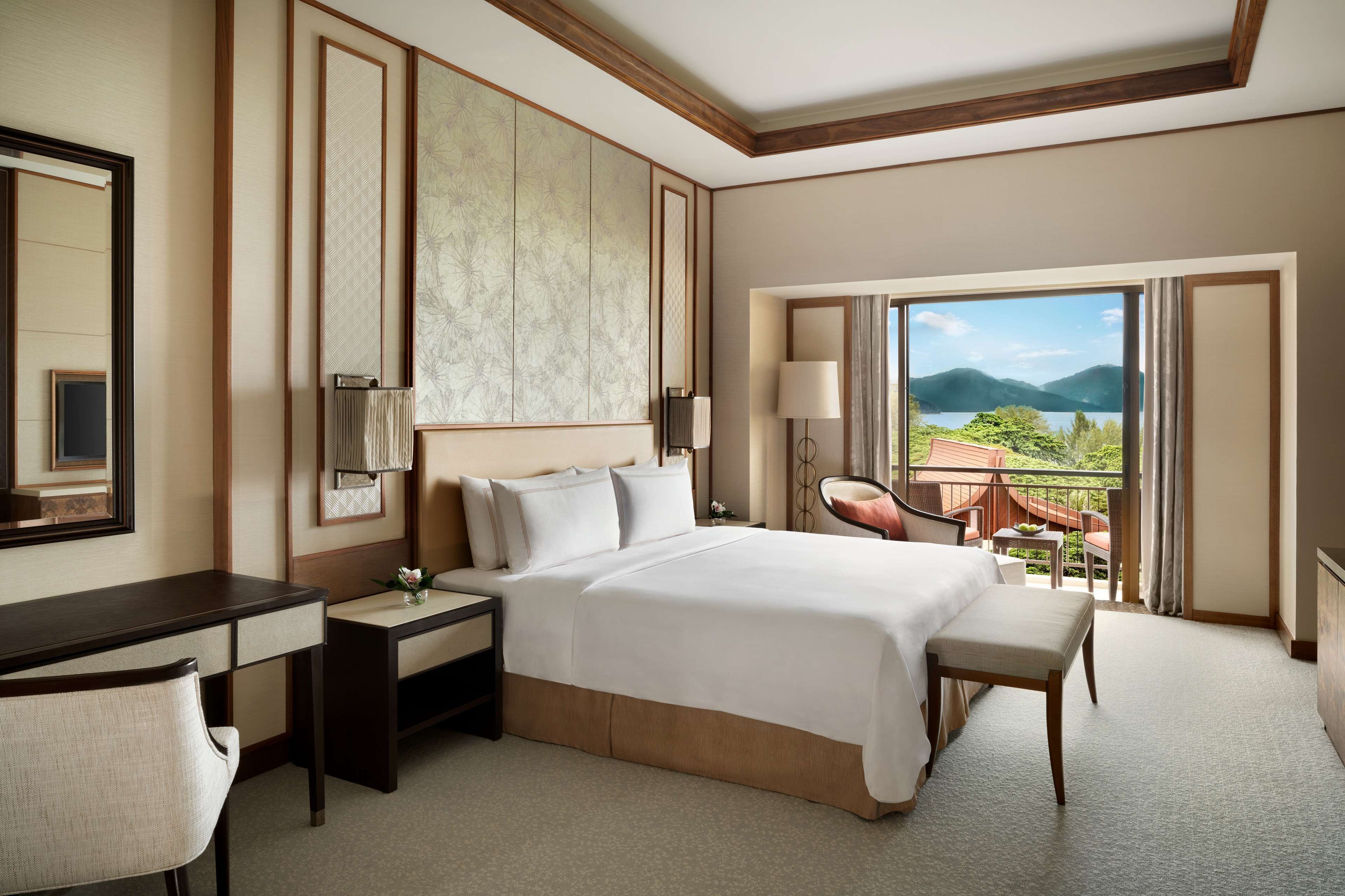 A hotel room with a large bed, a desk, a chair, a nightstand, a lamp, and a view of mountains and a lake.