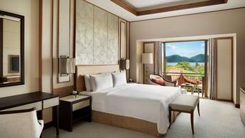 A hotel room with a large bed, a desk, a chair, a nightstand, a lamp, and a view of mountains and a lake.
